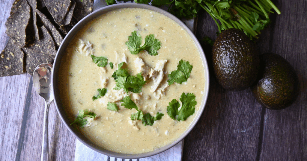 A bowl of creamy chicken soup garnished with fresh cilantro sits on a wooden table next to two avocados, sprigs of cilantro, and several dark tortilla chips. A spoon rests beside the bowl.