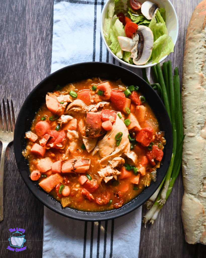 A bowl of chunky chicken and vegetable stew sits on a striped cloth, next to fresh green onions, a baguette, and a bowl of salad with lettuce, mushrooms, and tomatoes.
