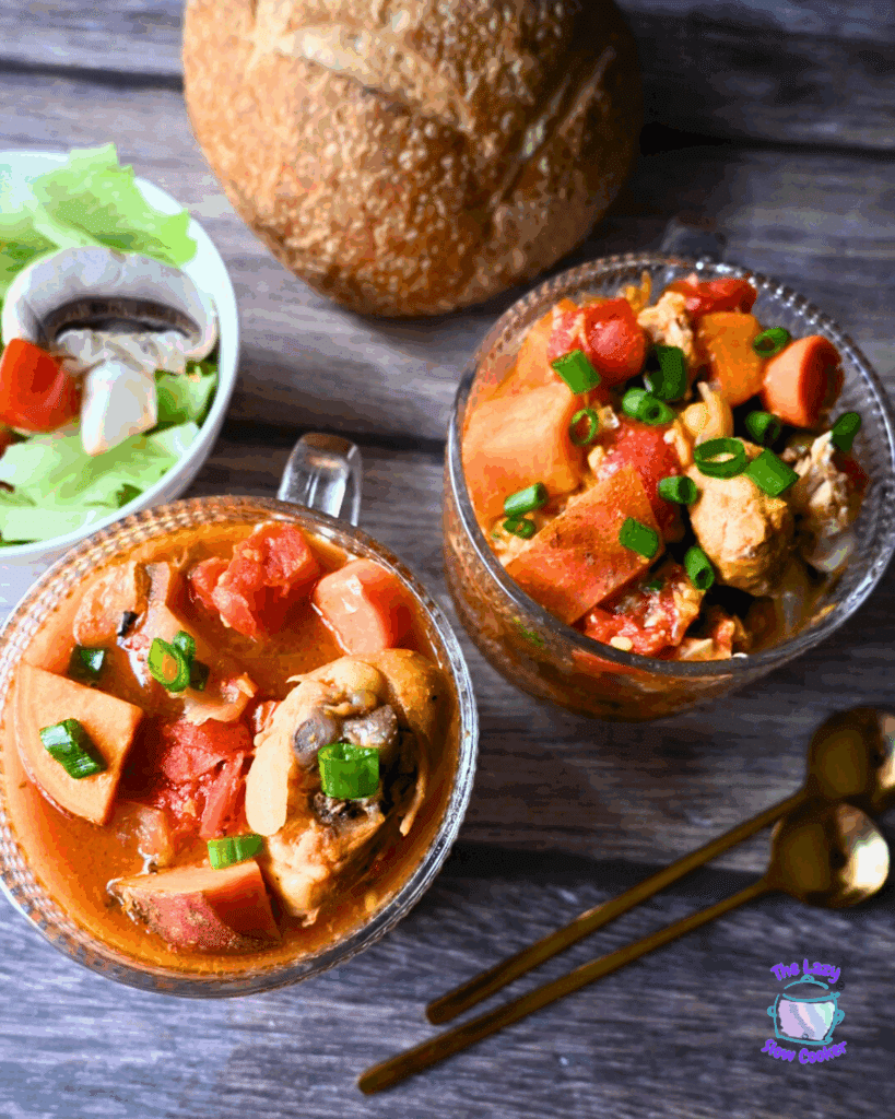 Two glass mugs filled with chunky chicken and vegetable stew, topped with chopped green onions, sit on a wooden surface next to a round loaf of bread, a salad bowl, and two golden spoons.