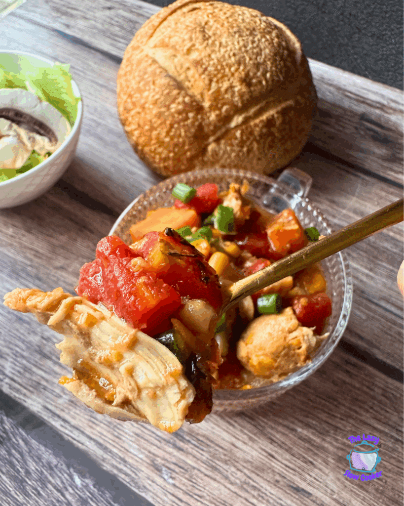 A spoonful of chunky chicken and tomato stew is held above a bowl, with a round loaf of crusty bread and a small salad in the background, all set on a wooden surface.