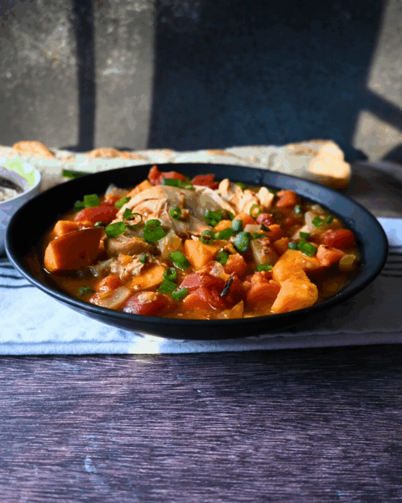 A black bowl filled with a hearty stew featuring chunks of sweet potato, tomatoes, chicken, and green onions sits on a table with a breadstick and napkin in the background.