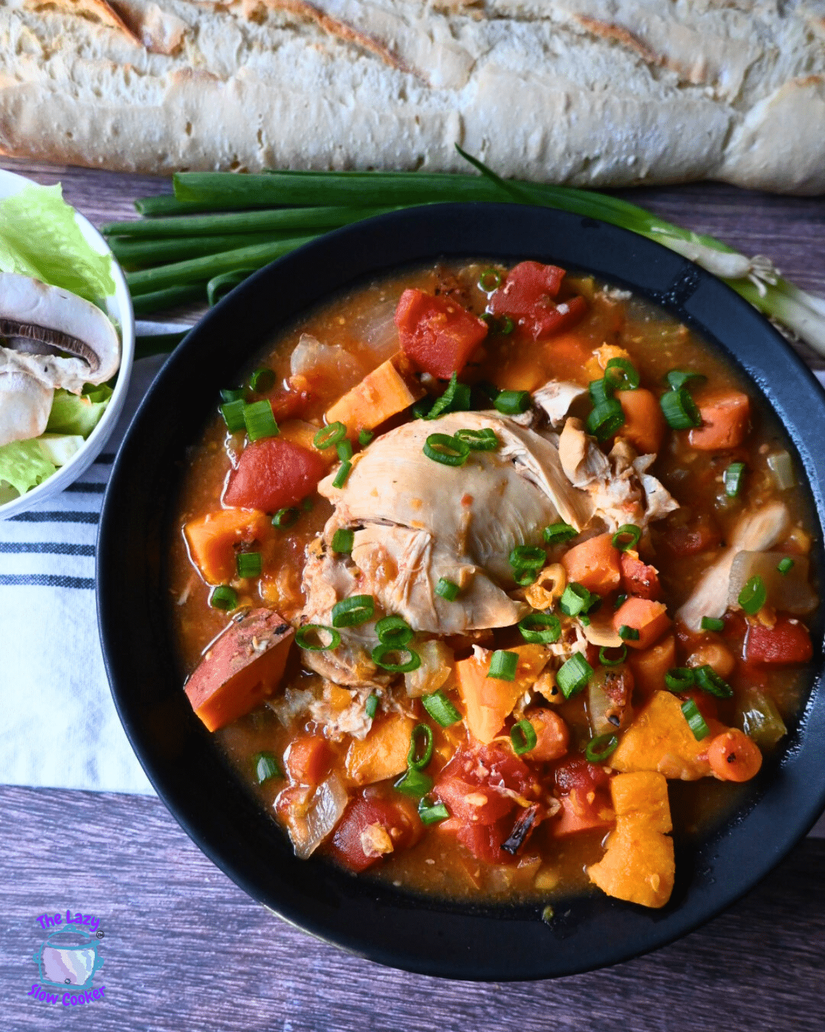 A bowl of chicken stew with sweet potatoes, tomatoes, and green onions, garnished with chopped green onions. A baguette, lettuce, mushrooms, and green onions are in the background on a wooden surface.