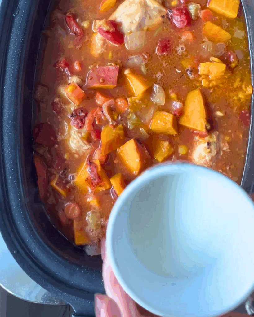 A slow cooker filled with a chunky stew containing chicken, tomatoes, and sweet potatoes. A hand is holding a small white bowl above the cooker, ready to scoop some stew.