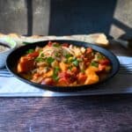 A black bowl filled with a hearty stew featuring chunks of sweet potato, tomatoes, chicken, and green onions sits on a table with a breadstick and napkin in the background.