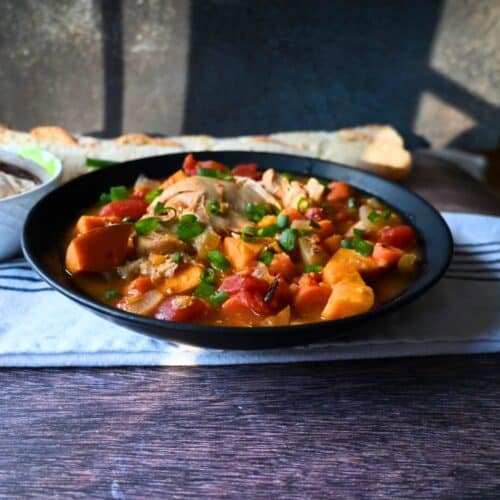A black bowl filled with a hearty stew featuring chunks of sweet potato, tomatoes, chicken, and green onions sits on a table with a breadstick and napkin in the background.