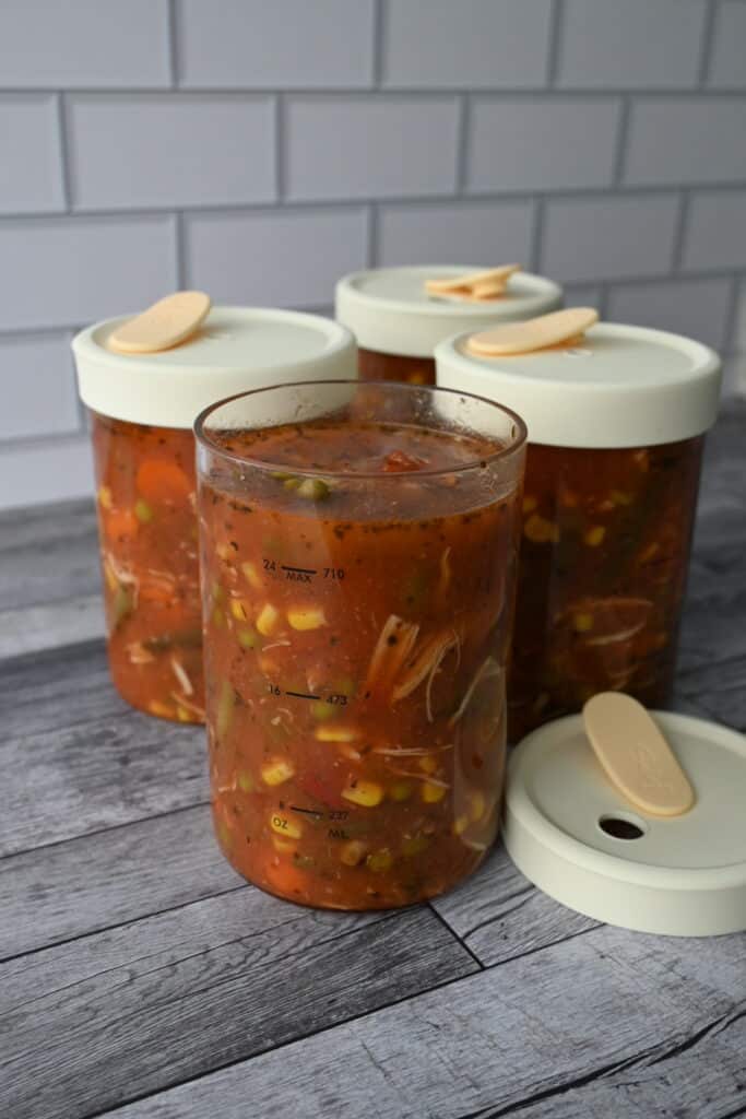 four glass containers of homemade vegetable soup in tomato broth are on a wood counter; three are sealed, with one lid set aside.