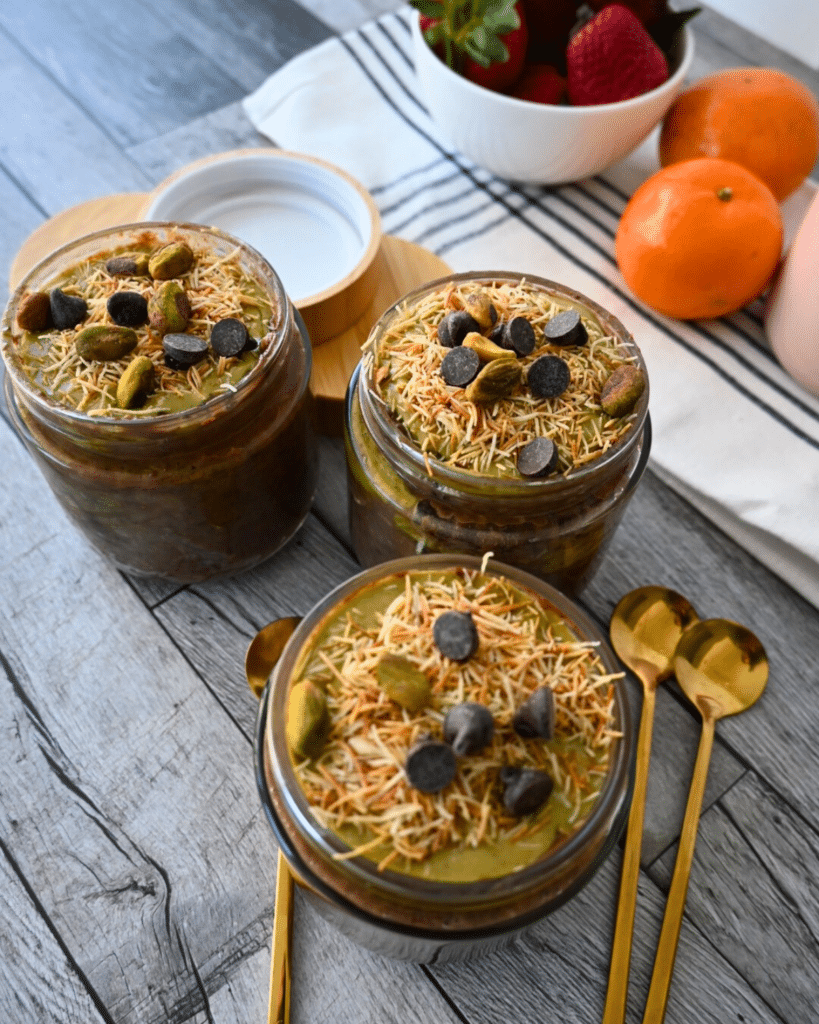 Three jars of slow cooker Dubai chocolate oatmeal with gold spoons laying on the side with fruit and a coffee cups