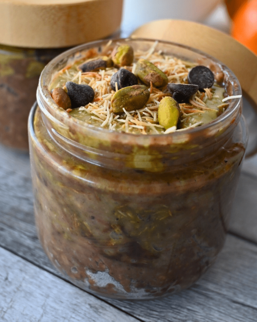 A jar with chocolate oats, pistachio butter, toasted kataifi and chocolate chips sits with a gold spoon. Text reads "Slow Cooker Dubai Chocolate Oats." Fruit and a QR code are in the background.