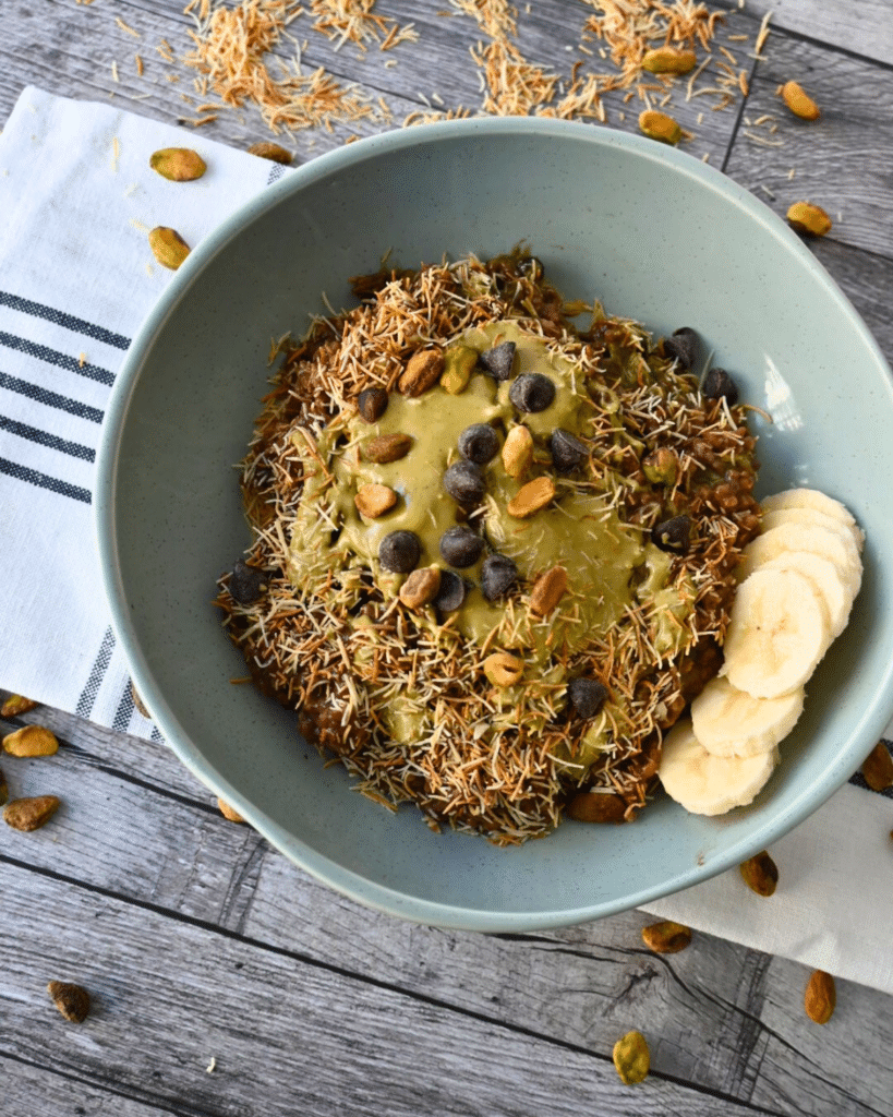 A blue bowl of oatmeal with kataifi, chocolate chips, nuts, nut butter, and banana slices on a striped cloth atop a wood table.