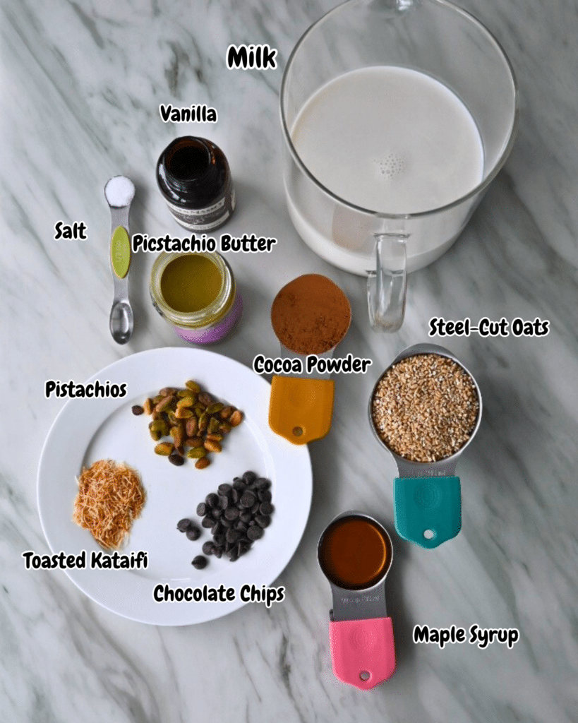 Labeled bowls and cups of ingredients for slow cooker Dubai chocolate oats are neatly arranged on a marble countertop. including pistachio butter, cocoa powder, and steel cut oats.