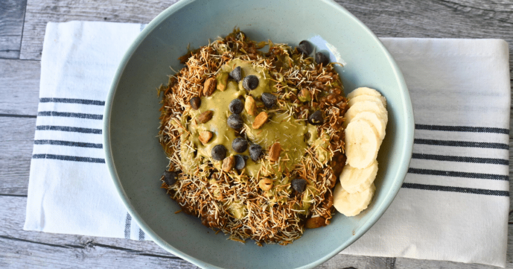 A blue bowl of oatmeal with kataifi, chocolate chips, nuts, nut butter, and banana slices on a striped cloth atop a wood table.