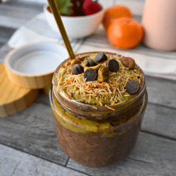 A jar of green smoothie topped with coconut, pistachios, and chocolate chips sits on a wooden surface with gold spoon and bamboo-lid jar.