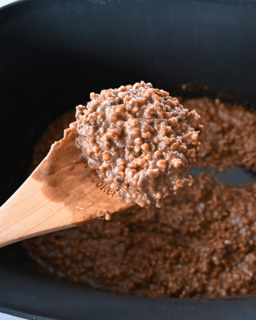 A wooden spoon lifts a serving of creamy chocolate oatmeal over a pot filled with more oatmeal, likely made in a slow cooker.