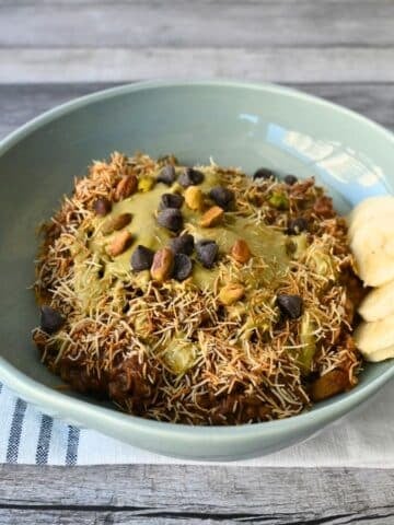 A blue bowl of oatmeal with kataifi, chocolate chips, nuts, nut butter, and banana slices on a striped cloth atop a wood table.