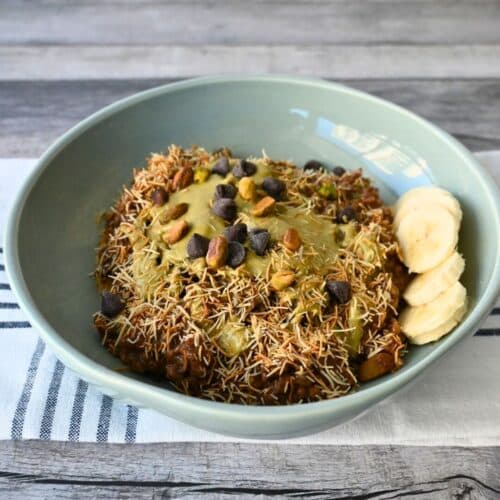 A blue bowl of oatmeal with kataifi, chocolate chips, nuts, nut butter, and banana slices on a striped cloth atop a wood table.