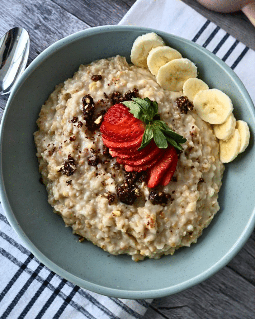 A bowl of steel-cut oats with bananas, strawberries, and raisins rests on a striped napkin beside a spoon. Text: Foolproof Slow Cooker Oats.