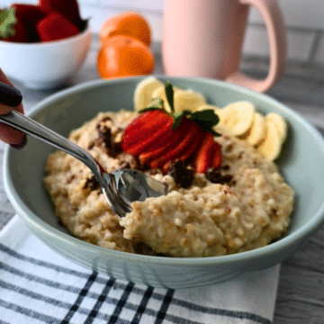 A hand holds a spoon in a bowl of oatmeal with strawberries, banana slices, and chocolate shavings on striped cloth; fruit and mug nearby.