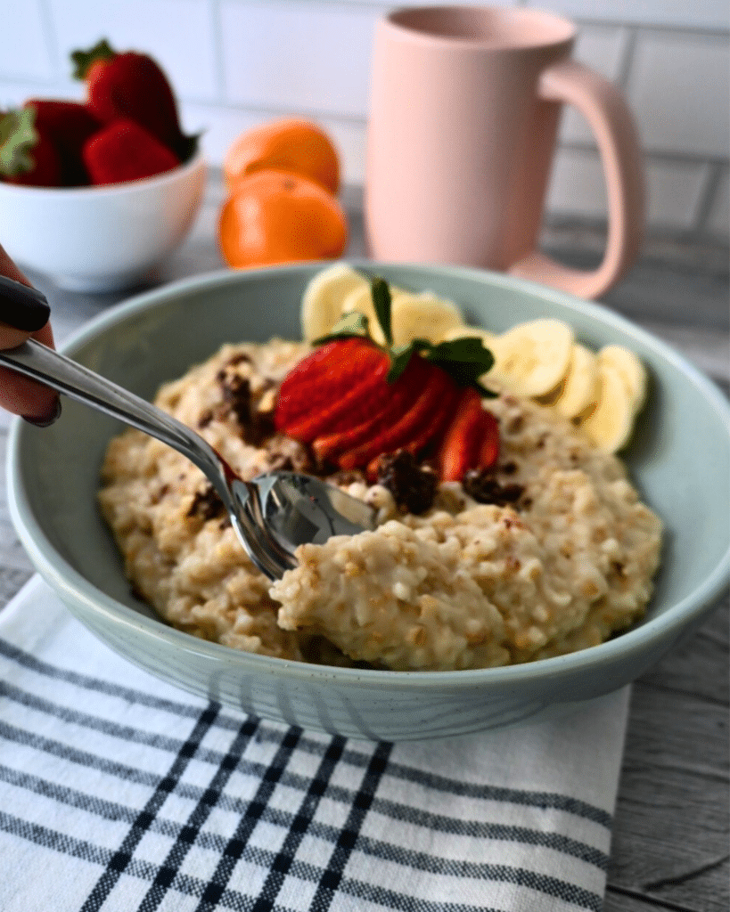 A hand holds a spoon in a bowl of oatmeal with strawberries, banana slices, and chocolate shavings on striped cloth; fruit and mug nearby.