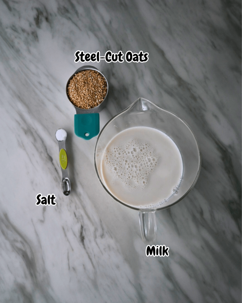 On a marble counter, labeled cups of milk and steel-cut oats sit beside a measuring spoon of salt for slow cooker oatmeal.