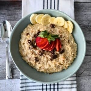 A bowl of steel-cut oats with bananas, strawberries, and raisins rests on a striped napkin beside a spoon.
