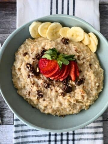 A bowl of steel-cut oats with bananas, strawberries, and raisins rests on a striped napkin beside a spoon.