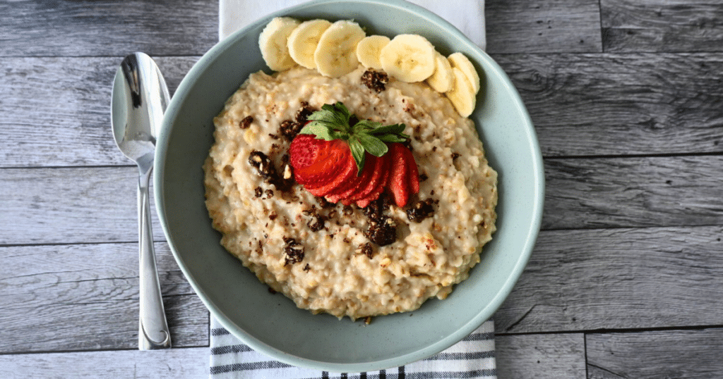 A bowl of steel-cut oats with bananas, strawberries, and raisins rests on a striped napkin beside a spoon.