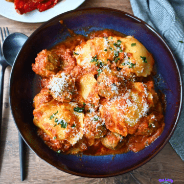A blue bowl of gnocchi and meatballs in tomato sauce with cheese and herbs, next to roasted red peppers and cutlery on a wood table.