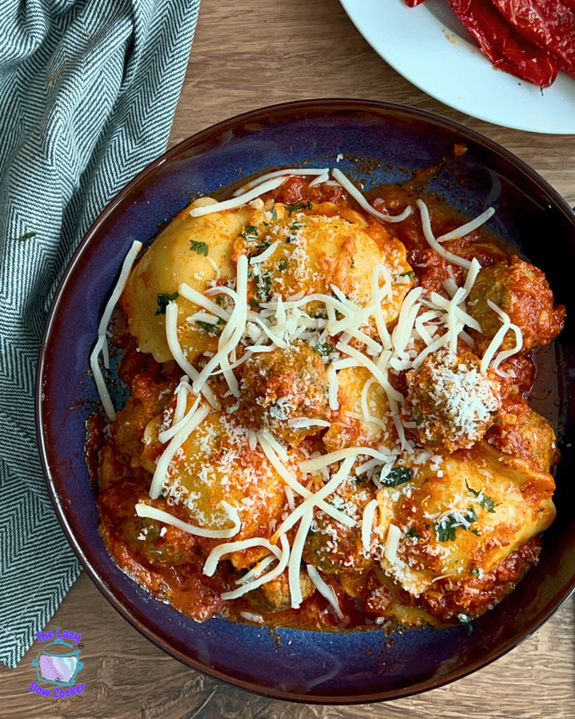 A blue bowl of ravioli in marinara sauce, topped with cheese and parsley, sits on wood beside a gray napkin and dried red peppers.