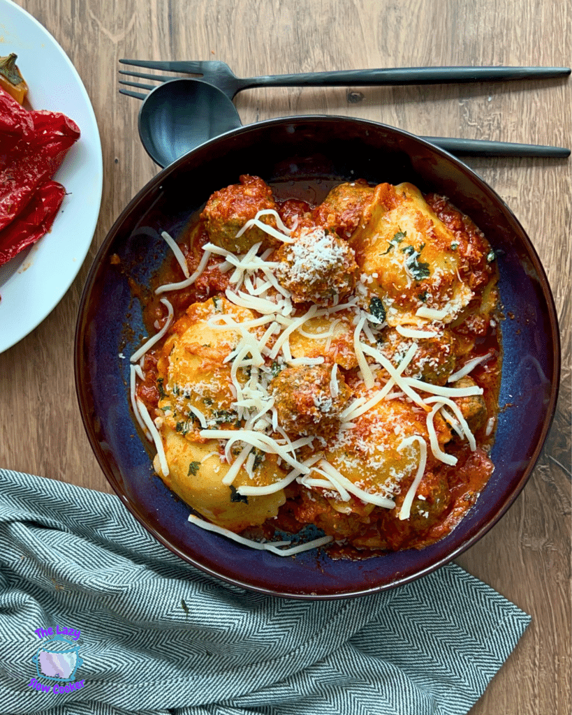 A bowl of ravioli with tomato sauce, cheese, and parsley on a wooden table next to red peppers, utensils, and a gray napkin.