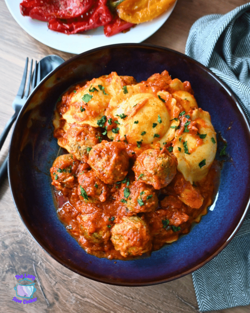 Meatballs in tomato sauce and mashed potatoes with herbs in a blue bowl, with roasted peppers and cutlery in the background.
