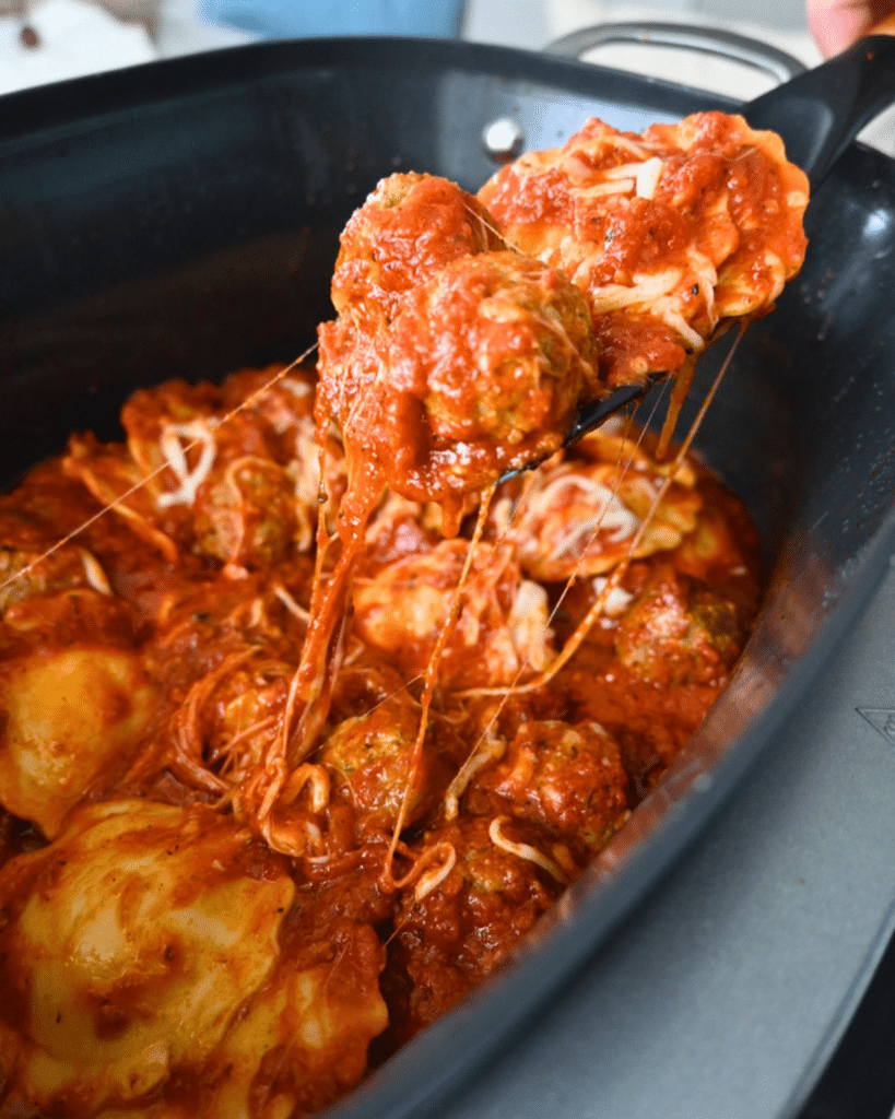 A slow cooker with cheesy ravioli and meatballs in tomato sauce, as a spoon lifts out a portion with melted cheese stretching.