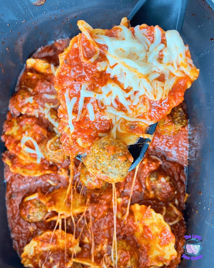 A spoon lifts cheesy ravioli and a meatball from a slow cooker, with stretchy melted cheese and more ravioli in rich tomato sauce below.