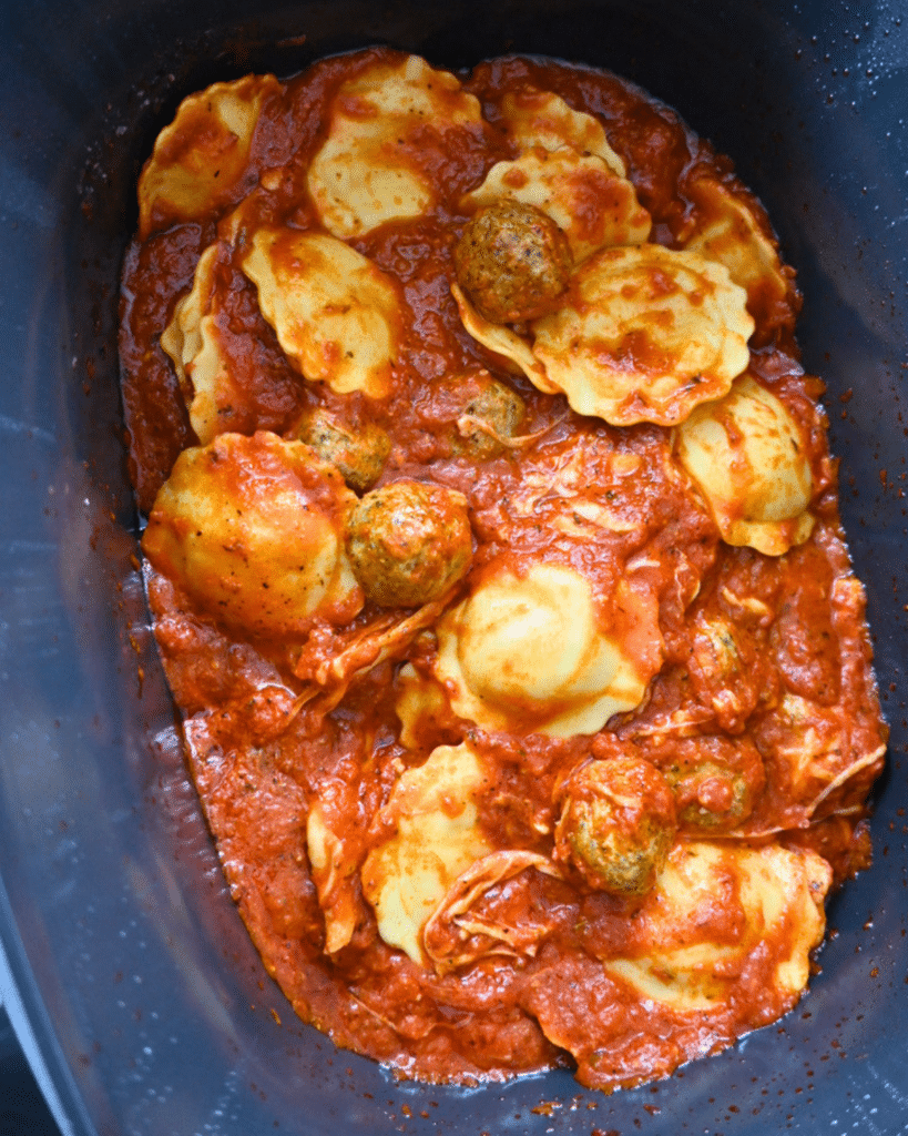 A slow cooker filled with cheese ravioli and meatballs smothered in marinara sauce, all simmering together.