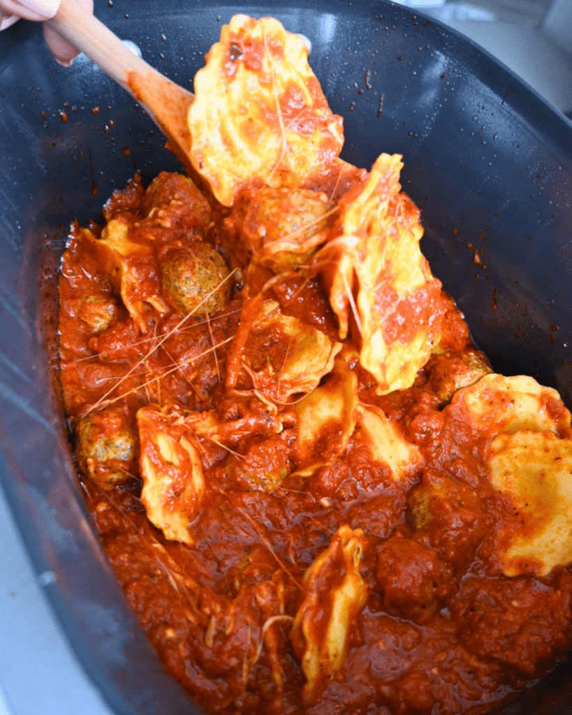 A wooden spoon scoops cheesy ravioli and meatballs coated in tomato sauce out of a black slow cooker for serving.