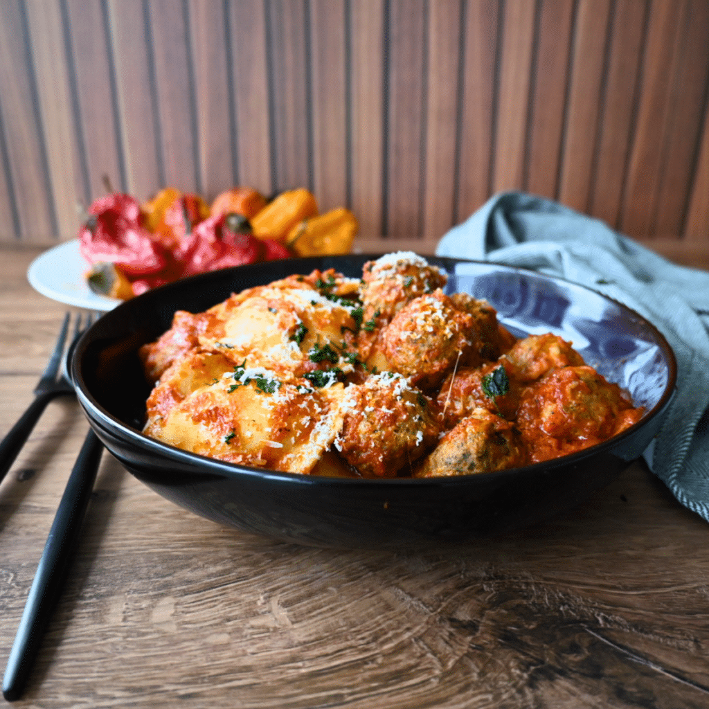 Gnocchi and meatballs in tomato sauce, topped with cheese and herbs, served in a black bowl on a wooden table with sides nearby.
