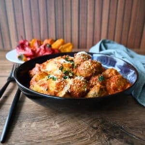 A black bowl of slow cooker meatballs in tomato sauce, topped with cheese and herbs, sits on a wooden table beside roasted peppers.