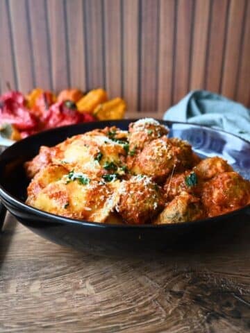 A black bowl of slow cooker meatballs in tomato sauce, topped with cheese and herbs, sits on a wooden table beside roasted peppers.