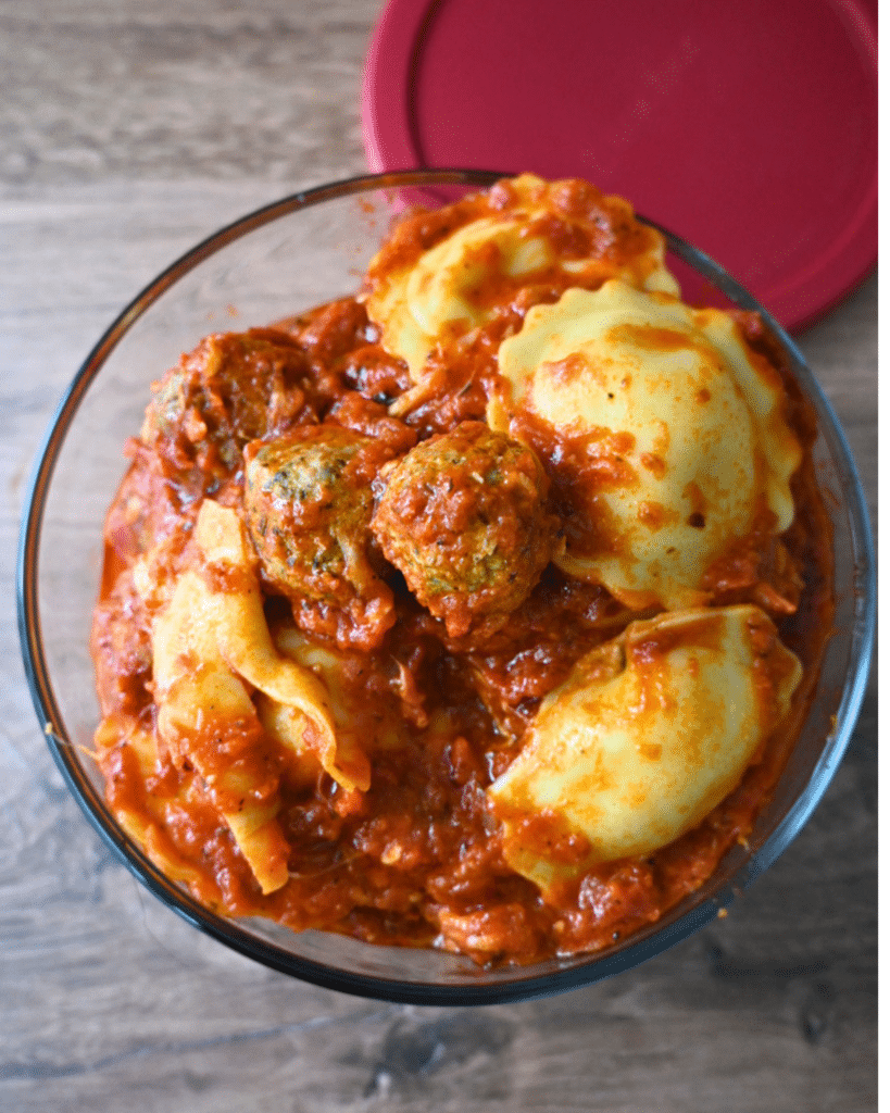 A glass bowl with leftover ravioli and meatballs in tomato sauce rests on a wooden table, with a red lid set beside the bowl.