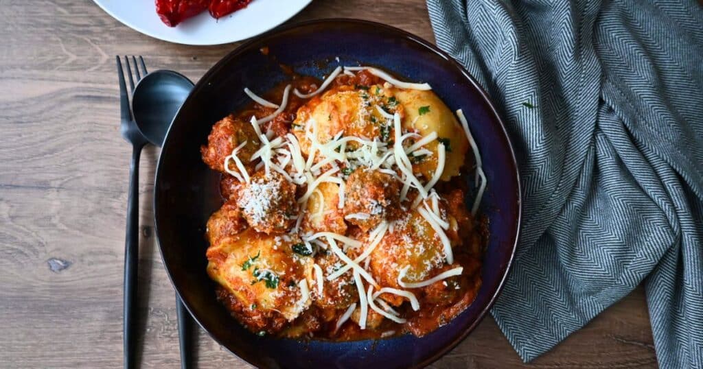A bowl of ravioli and meatballs with melted cheese and herbs sits on a wooden table, fork, spoon, and gray napkin beside it.