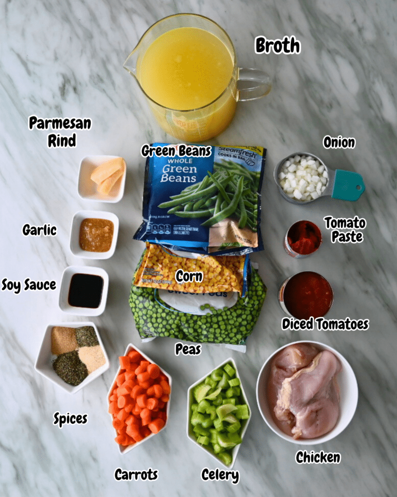 Ingredients for slow cooker chicken soup, each labeled and arranged on a marble countertop in an organized overhead layout. including chyicken, spices, peas, carrots, celery, corn, onion, green beans, diced tomatoes, tomato paste, garlic, soy sauce, Parmesan rind and broth