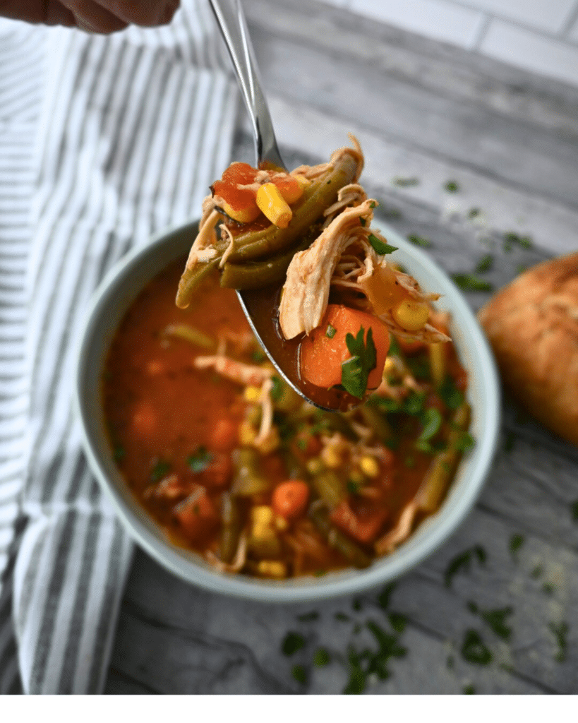 A spoonful of slow cooker chicken vegetable soup with shredded chicken and veggies is held over a bowl, with bread and a towel nearby.