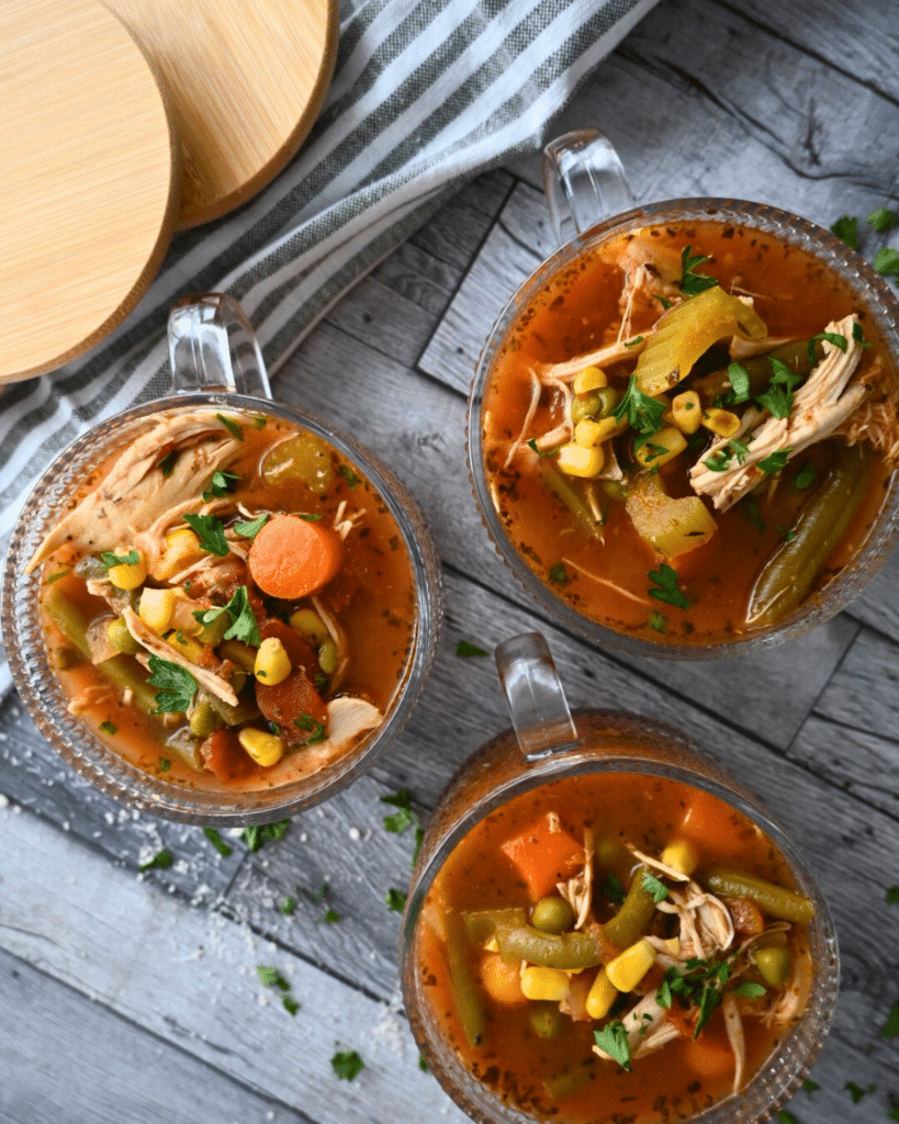 Three glass mugs of chunky vegetable soup with parsley and celery garnish sit on a wooden table, parsley scattered around