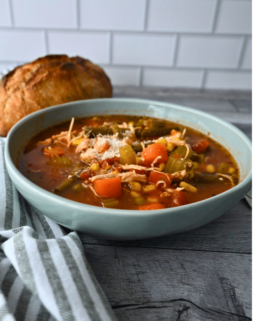 A bowl of chicken and vegetable soup topped with cheese sits on a wooden table beside bread, with a striped napkin and tiled wall.