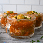 Three glass mugs of chunky vegetable soup with parsley and celery garnish sit on a wooden table, parsley scattered around, white tile behind.