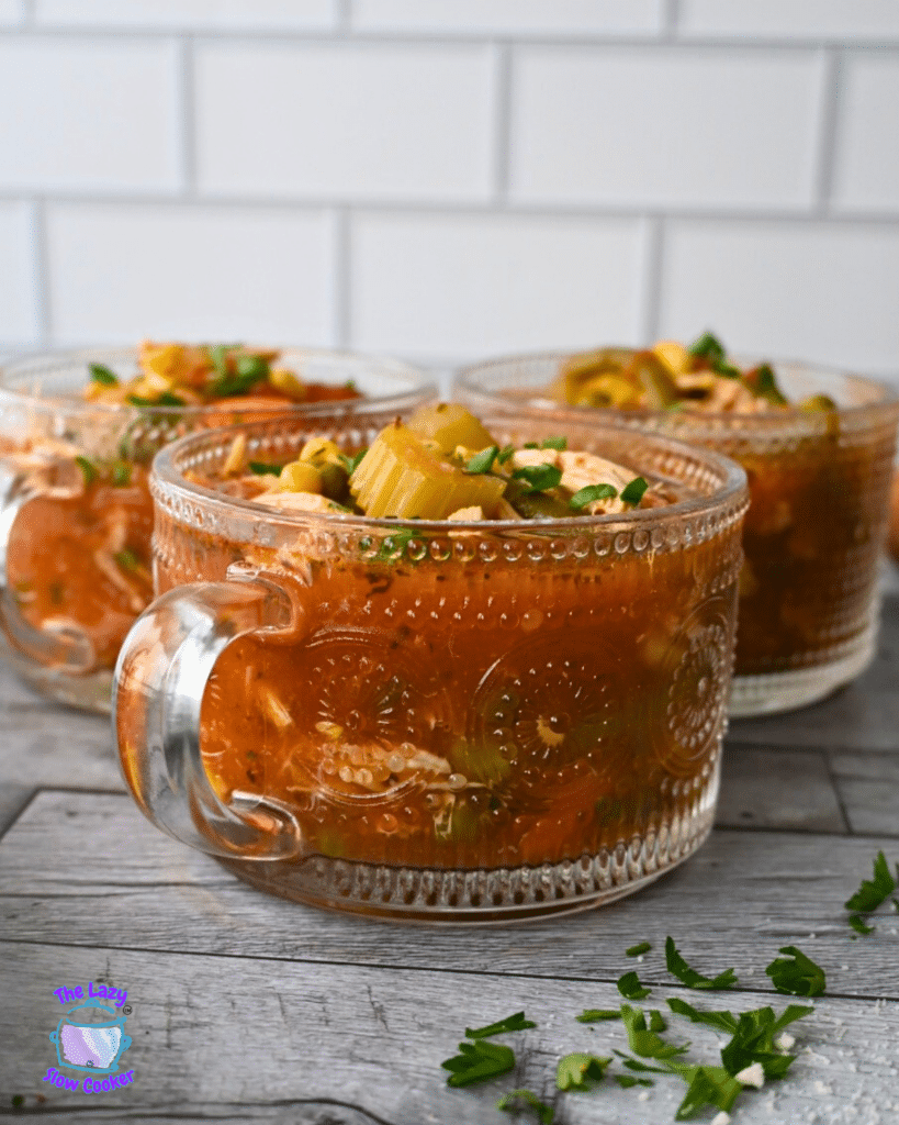 Three glass mugs of chunky vegetable soup with parsley and celery garnish sit on a wooden table, parsley scattered around, white tile behind.