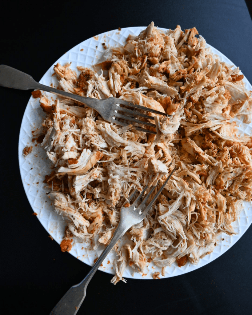Shredded chicken cooked with spices sits on a white plate, two forks resting on top, set against a dark background.