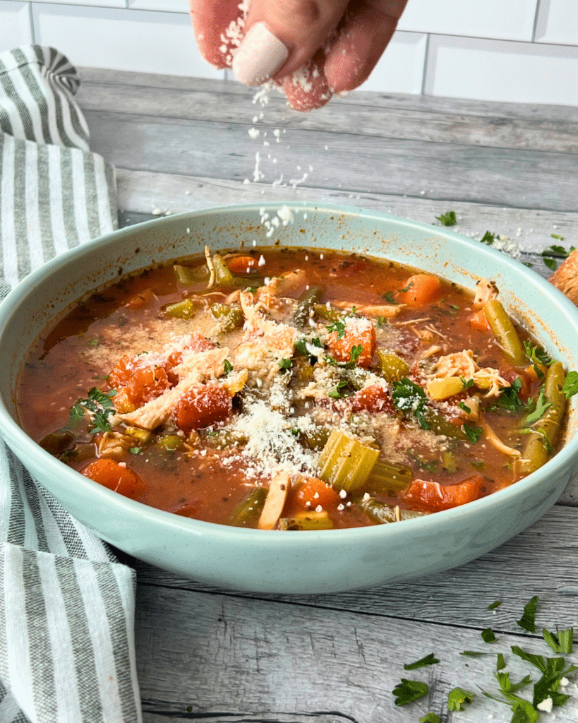 A hand sprinkles grated cheese over chicken and vegetable soup topped with parsley in a blue bowl on a wooden table.