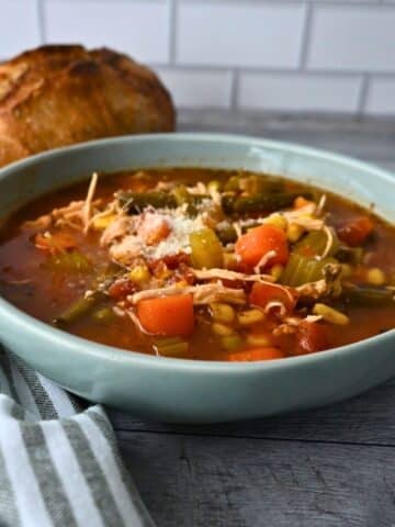 A bowl of chicken and vegetable soup topped with cheese sits on a wooden table beside bread, with a striped napkin and tiled wall.