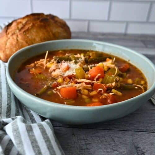 A bowl of chicken and vegetable soup topped with cheese sits on a wooden table beside bread, with a striped napkin and tiled wall.