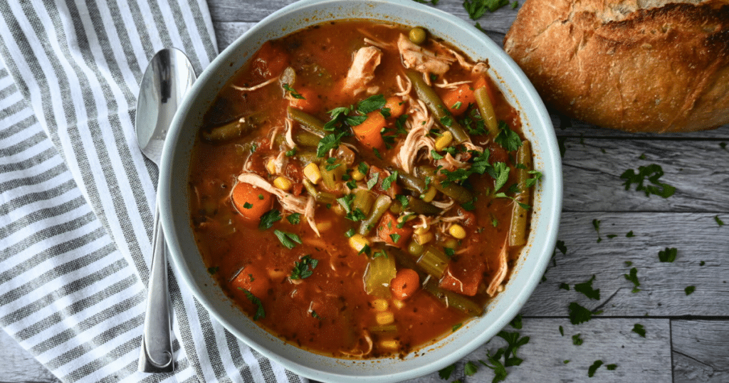 A bowl of chicken vegetable soup with carrots, green beans, corn, and shredded chicken in tomato broth sits by a loaf of bread.